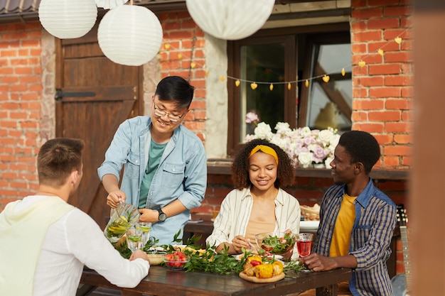 Groupe multiethnique d'amis assis à une table en bois tout en appréciant le dîner en plein air à la fête d'été