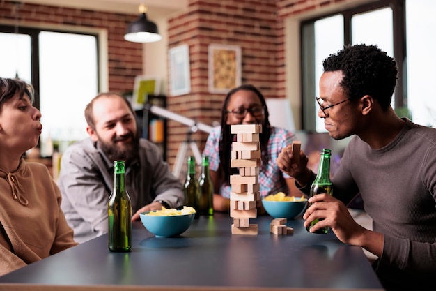 Groupe multiethnique d'amis assis à table dans le salon tout en jouant ensemble à des jeux de société. Diverses personnes assises à la maison tout en profitant d'un jeu de société avec des blocs de bois et une structure de tour.