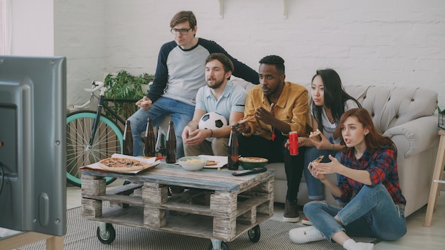 Groupe multiethnique d'amis fans de sport regardant le championnat de football à la télévision ensemble mangeant de la pizza et buvant de la bière à la maison