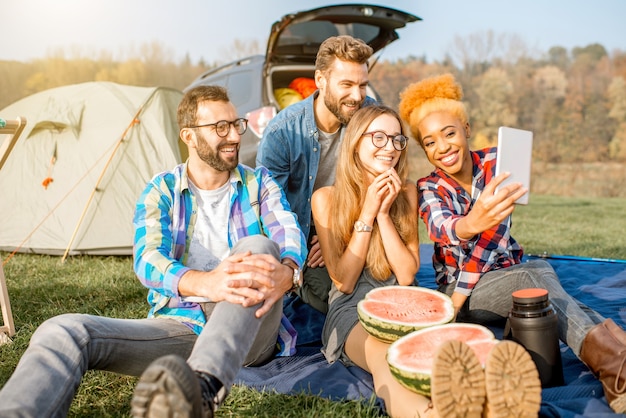 Groupe multiethnique d'amis habillés avec désinvolture s'amusant à faire une photo de selfie ensemble pendant les loisirs de plein air avec tente, voiture et équipement de randonnée près du lac