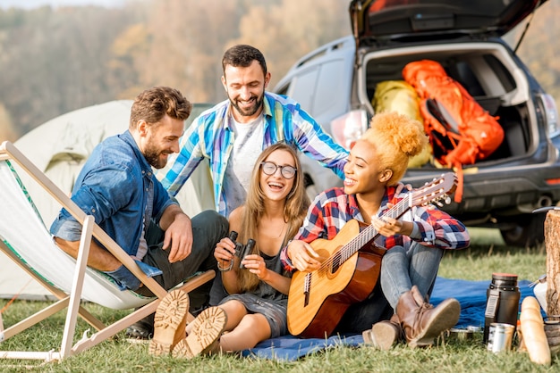 Groupe multiethnique d'amis habillés avec désinvolture s'amusant à jouer de la guitare pendant les loisirs de plein air avec tente près du lac