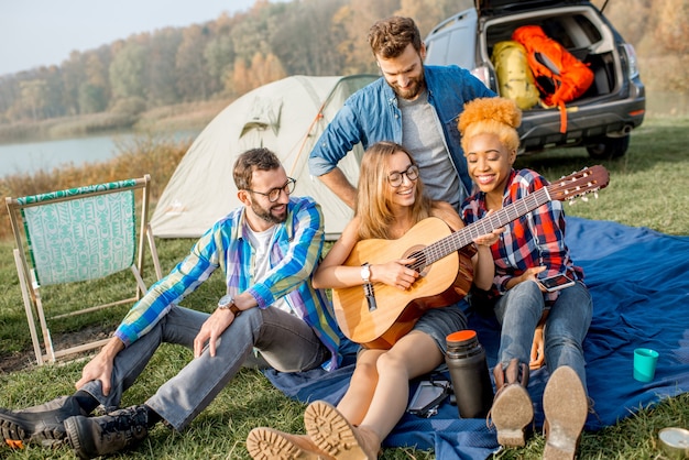 Groupe multiethnique d'amis habillés avec désinvolture s'amusant à jouer de la guitare pendant les loisirs de plein air avec tente, voiture et équipement de randonnée près du lac