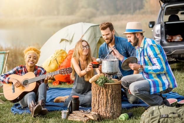 Groupe multiethnique d'amis habillés avec désinvolture en train de pique-niquer, de cuisiner une soupe avec un chaudron, de jouer de la guitare pendant les loisirs de plein air près du lac