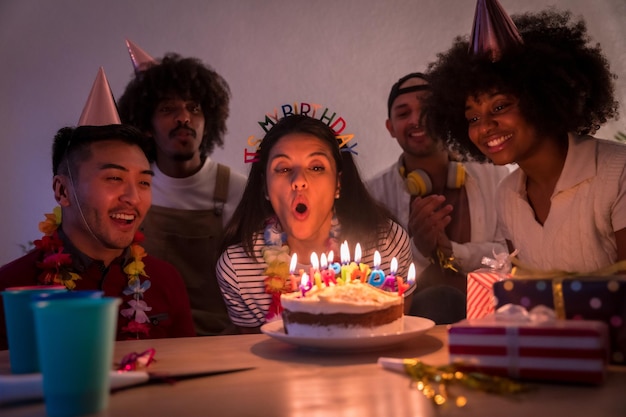 Groupe multiethnique d'amis lors d'une fête d'anniversaire sur le canapé à la maison avec un gâteau et des cadeaux soufflant les bougies avec les lumières éteintes