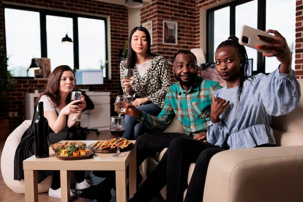 Groupe multiethnique d'amis prenant une photo de selfie ensemble lors d'une fête du vin dans le salon. Heureux adultes souriants à la maison profitant du temps ensemble tout en buvant des boissons alcoolisées et en prenant des photos