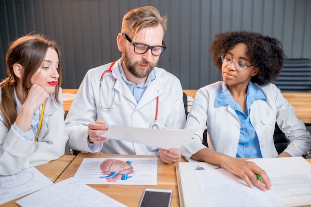 Groupe multiethnique d'étudiants en médecine en uniforme discutant assis ensemble au bureau avec différents trucs médicaux en classe