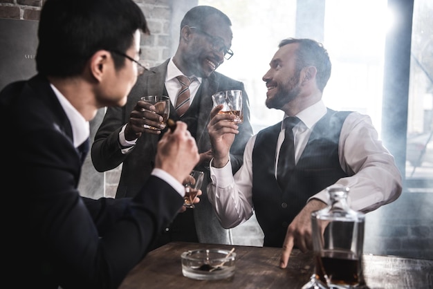 Groupe multiethnique d'hommes d'affaires fumant et buvant du whisky à l'intérieur, équipe commerciale multiculturelle