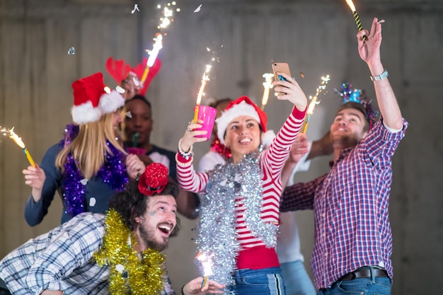 groupe multiethnique de jeunes gens d'affaires occasionnels heureux prenant une photo de selfie tout en dansant avec des cierges magiques et en s'amusant pendant la fête du nouvel an devant un mur de béton au nouveau bureau de démarrage