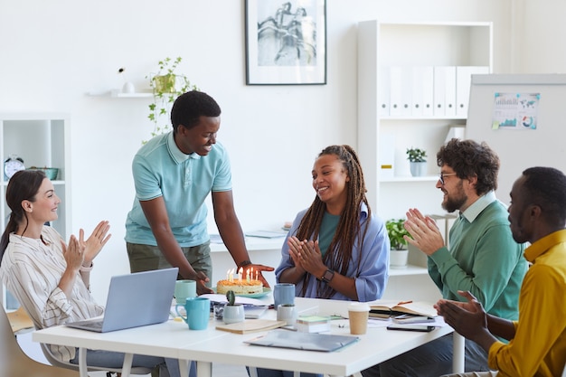 Groupe multiethnique de personnes célébrant l'anniversaire au bureau, se concentrer sur un jeune homme souriant apportant un gâteau à une femme afro-américaine