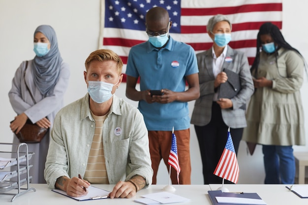 Groupe multiethnique de personnes debout en ligne et portant des masques au bureau de vote le jour de l'élection, se concentrer sur le jeune homme lors de l'inscription pour voter