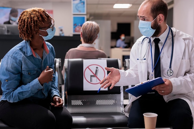 Groupe multiethnique de personnes effectuant une visite de contrôle dans la salle d'attente du hall d'accueil de l'hôpital. Médecin généraliste consultant une femme avec un masque anti-virus préventif dans un établissement médical.