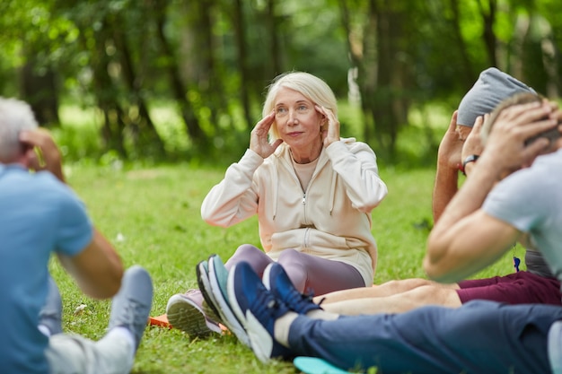 Groupe de personnes âgées modernes réunis dans le parc assis sur des nattes sur l'herbe faisant étirement du cou exercice de détente