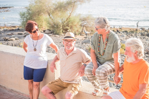 Groupe de personnes âgées et de personnes mûres assis à la plage sur un banc - amitié heureuse avec deux couples de retraités mariés parlant et s'amusant