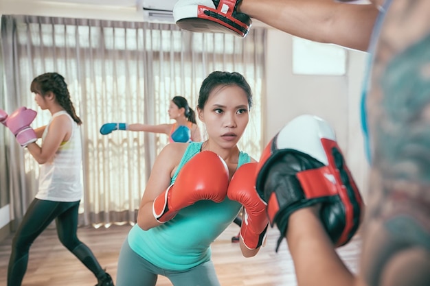 un groupe de personnes asiatiques rejoignent un club de boxe faisant des exercices de boxe dans la salle de sport. jeune femme sportive avec sa formation d'entraîneur personnel prête à pratiquer la pose de frappe et de poinçon avec l'entraîneur. filles sportives avec des gants.