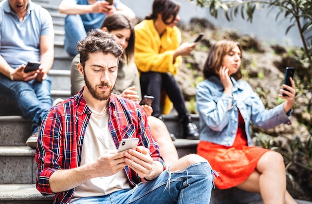 Groupe de personnes ennuyées utilisant un téléphone intelligent mobile assis dans la cour d'un collège urbain Jeunes amis accros aux appareils technologiques pour smartphones Concept technologique avec des étudiants toujours connectés sur un filtre vif et lumineux