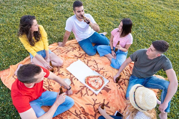 Groupe de personnes heureuses au parc mangeant de la pizza et buvant des bières en bouteille