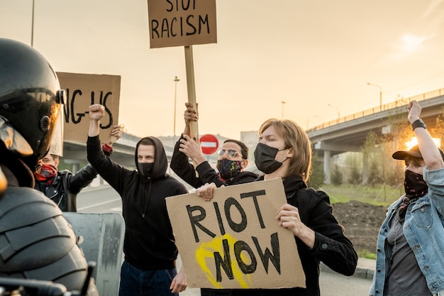 Groupe de personnes mécontentes portant des masques avec des pancartes revendiquant l'égalité des droits pour toutes les ethnies tout en criant contre la police anti-émeute sur la route