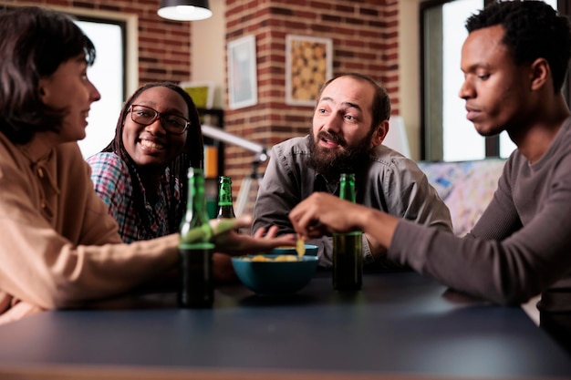 Groupe positif d'amis multiethniques assis à table tout en discutant et en s'amusant ensemble. Des personnes multiraciales à la maison dans le salon qui savourent des collations et des boissons tout en parlant.