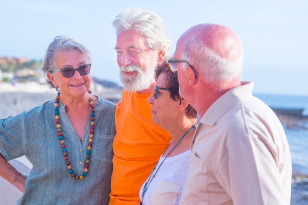 Groupe de quatre personnes âgées heureuses et personnes mûres parlant et s'amusant ensemble à la plage avec la mer en arrière-plan - amitié et relation cocncept