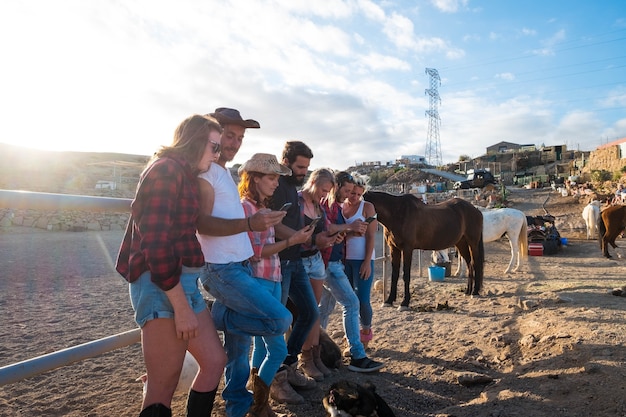 Groupe de sept personnes utilisant leur téléphone ensemble dans un ranch avec des chevaux en arrière-plan - personnes en ligne et réseaux ou réseaux sociaux