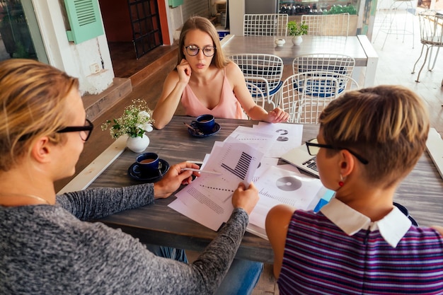 Un groupe de trois étudiants indépendants élégants entrepreneurs à la mode bien habillés travaillant assis à table avec un ordinateur portable dans un café d'été en plein air indépendant, travail à distance à distance, surfeur.