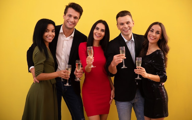 Un groupe de trois femmes et deux hommes, vêtus de tenues étonnantes et arborant des sourires sincères, tintent avec des verres pleins de champagne, faisant une fête de Noël ensemble.