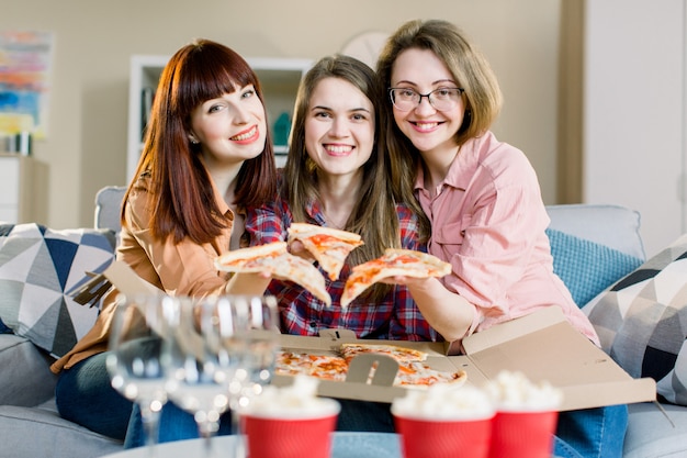 Groupe de trois jeunes filles amis manger de la pizza pendant la fête à la maison. Groupe de jeunes femmes s'amusant ensemble. Femmes heureuses parler et rire tout en mangeant de la nourriture italienne et assis sur un canapé.