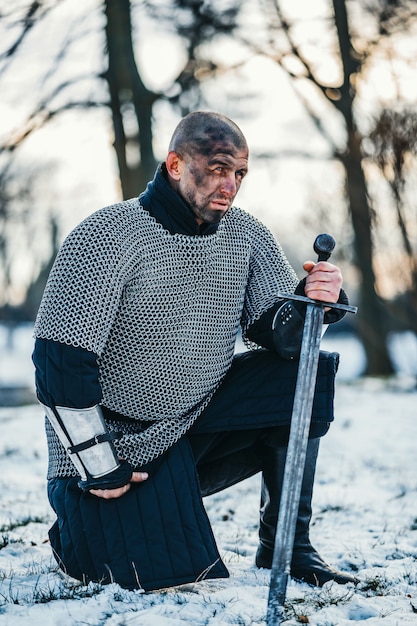 Un guerrier médiéval en cotte de mailles à genoux avec son épée à la main et le visage sale après la bataille.