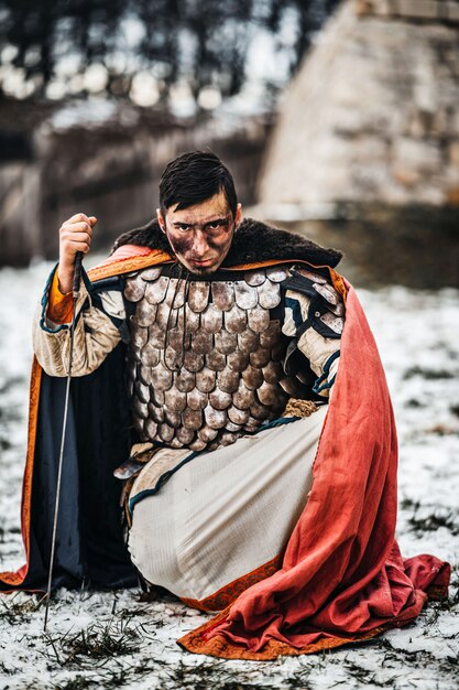 Un guerrier médiéval en cotte de mailles et manteau à genoux et tient sa main à l'épée après la bataille