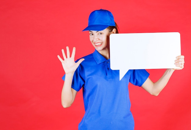 Guide féminin en uniforme bleu tenant un panneau d'information rectangulaire blanc et saluant les participants.