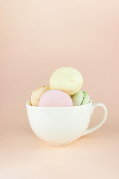 Les guimauves de biscuits à la meringue française ressemblent à des macarons dans une tasse à café en porcelaine blanche sur fond beige pastel