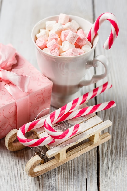 Guimauves blanches et roses dans une tasse, cannes de Noël et luge jouet sur table blanche.