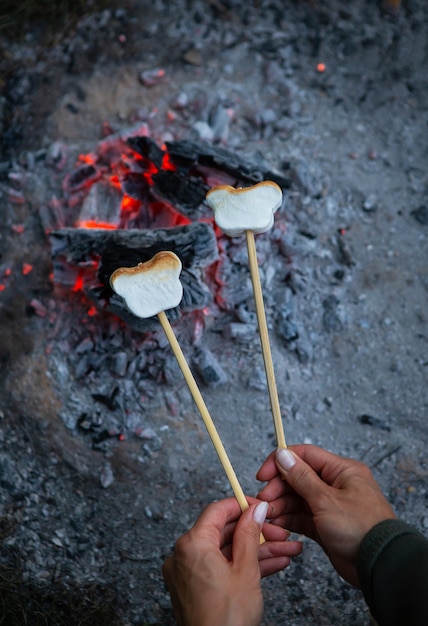 Les guimauves sont frites sur des brochettes sur un feu. Guimauves frites avec une flamme nue sur une brochette. Romance de feu de camp, soirée d'été. Vue d'en-haut.