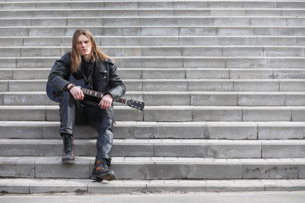 Guitariste rock sur les marches. Un musicien avec une guitare basse dans un costume en cuir. Metalist avec une guitare sur fond d'étape industrielle.