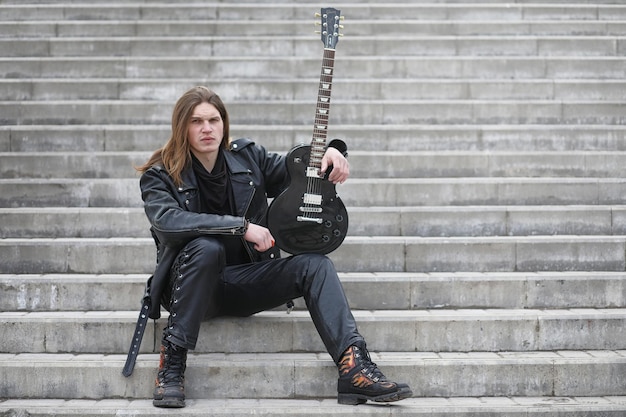 Guitariste rock sur les marches Un musicien avec une guitare basse dans un costume en cuir Métaliste avec une guitare sur fond de stepxA industriel