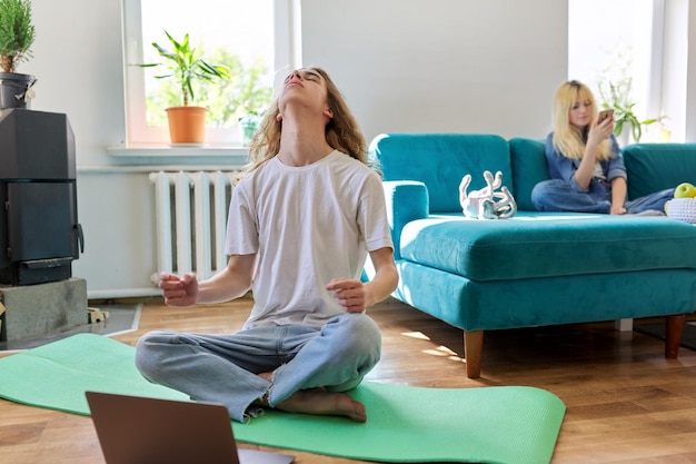 Guy adolescent assis en position du lotus sur un tapis de yoga à la maison sur le sol avec un ordinateur portable