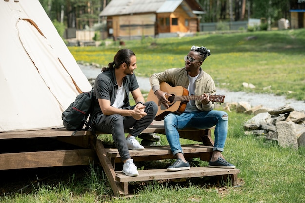 Guy assis sur l'étape et jouant de la guitare pour un ami pendant qu'ils passent du temps au festival de la campagne