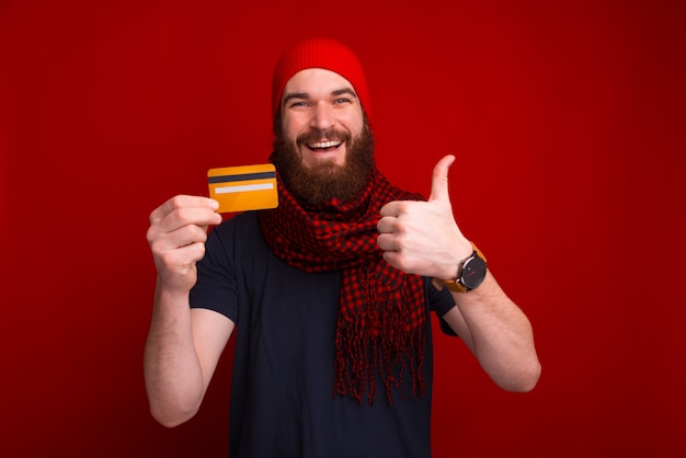 Guy barbu, en écharpe rouge et chapeau, montrant sa carte de crédit et son pouce, debout sur un espace rouge