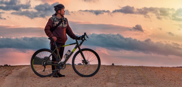 Guy dans un casque se dresse avec un vélo de gravier dans un champ de sable contre un ciel nuageux au coucher du soleil