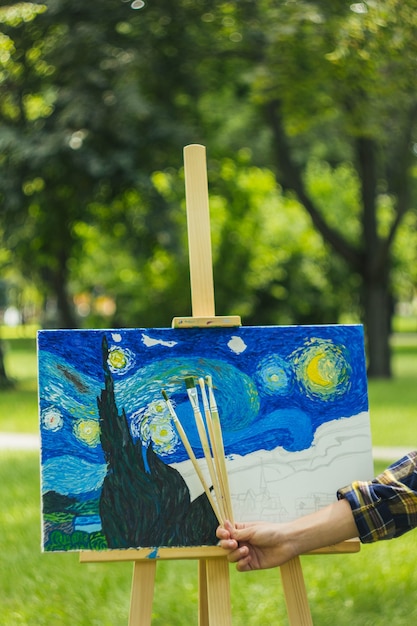 Guy dans un chapeau de paille est assis dans le parc devant un chevalet avec des peintures à l'huile