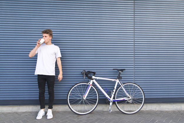 Guy dans un T-shirt blanc se tient à côté d'un vélo sur fond de mur et boit du café de tasse