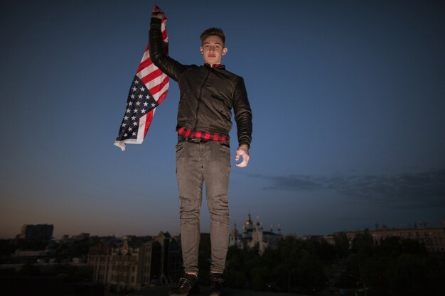 Guy avec drapeau volant américain dans la ville de nuit. Un fier patriote américain célèbre l'événement national. Un adolescent en vacances s'amuse à la fête de l'indépendance