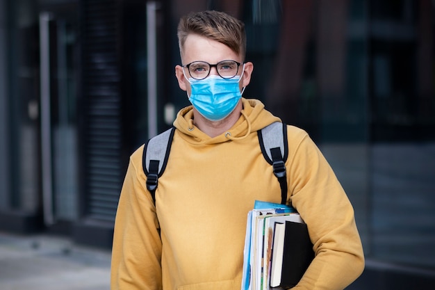 Guy étudiant, élève garçon, jeune homme dans un masque médical de protection et des lunettes sur le visage à l'extérieur de l'université avec des livres