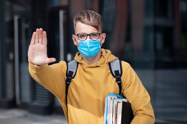 Guy étudiant, élève garçon, jeune homme en masque médical protecteur et lunettes sur le visage à l'extérieur de l'université avec des livres, des manuels scolaires montrent la paume, la main, n'arrêtez aucun signe. Virus, concept de coronavirus pandémique. Covid-19