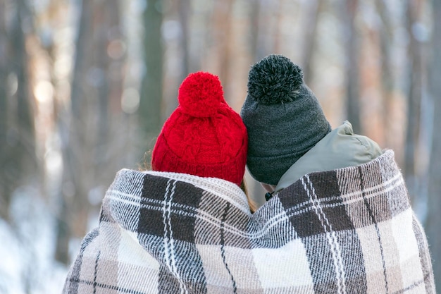 Guy et fille en bonnets tricotés avec des bubons en hiver dans le parc sont recouverts d'une couverture chaude. Vue arrière.