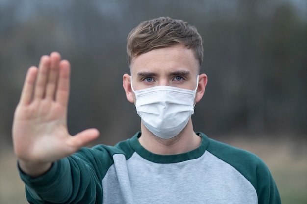 Guy, jeune homme dans un masque médical stérile protecteur sur son visage regarde la caméra à l'extérieur, sur la rue asiatique, paume, main, n'arrêtez aucun signe. Pollution atmosphérique, virus, concept de coronavirus pandémique. Covid-19