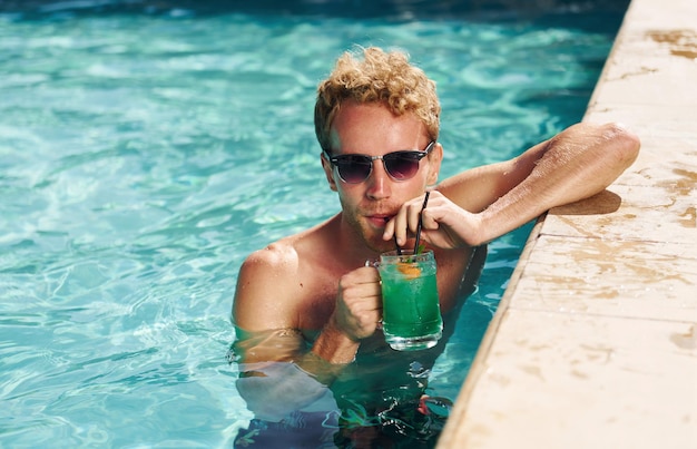 Guy en lunettes de soleil noires et avec cocktail est dans la piscine