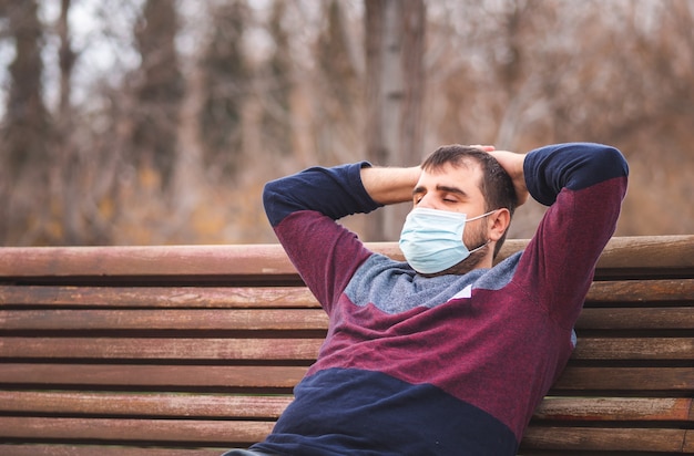 Guy avec masque de protection relaxant assis sur un banc de parc au coucher du soleil