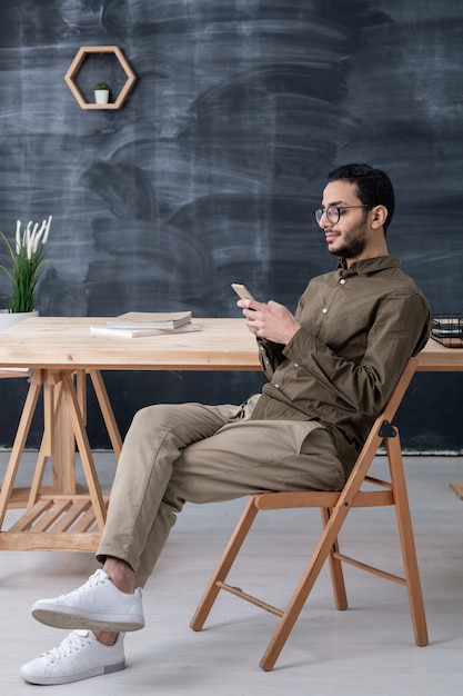 Guy sérieux assis sur une chaise par table en bois au bureau lors de la recherche de données dans le smartphone à la pause