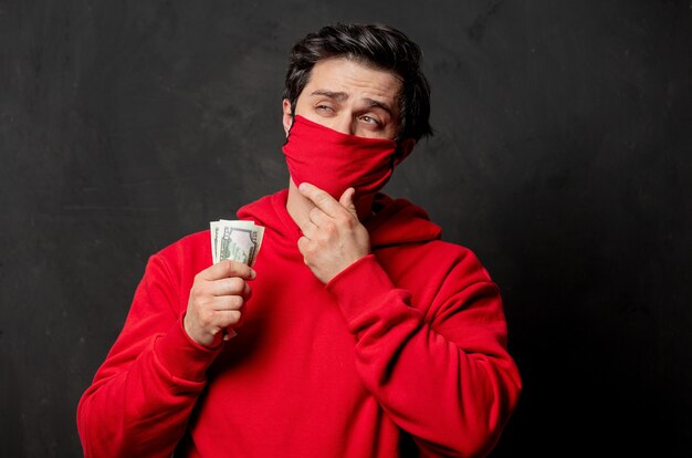 Guy en sweat-shirt rouge et masque facial tiennent de l'argent dans la main sur un mur sombre